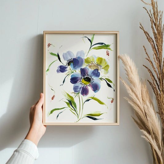 Hand holding a light wood frame containing a blue anemone print beside dried grasses.