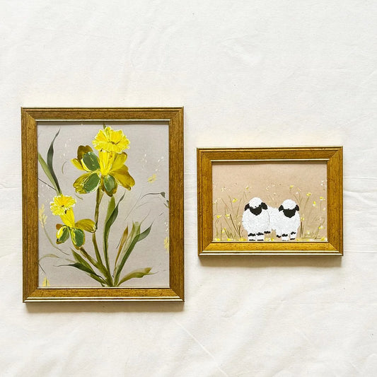 Two framed art prints hanging on a textured white wall. The larger left frame shows yellow flowers with green leaves on a light background, and the smaller right frame shows two white sheep with black faces standing in a field.