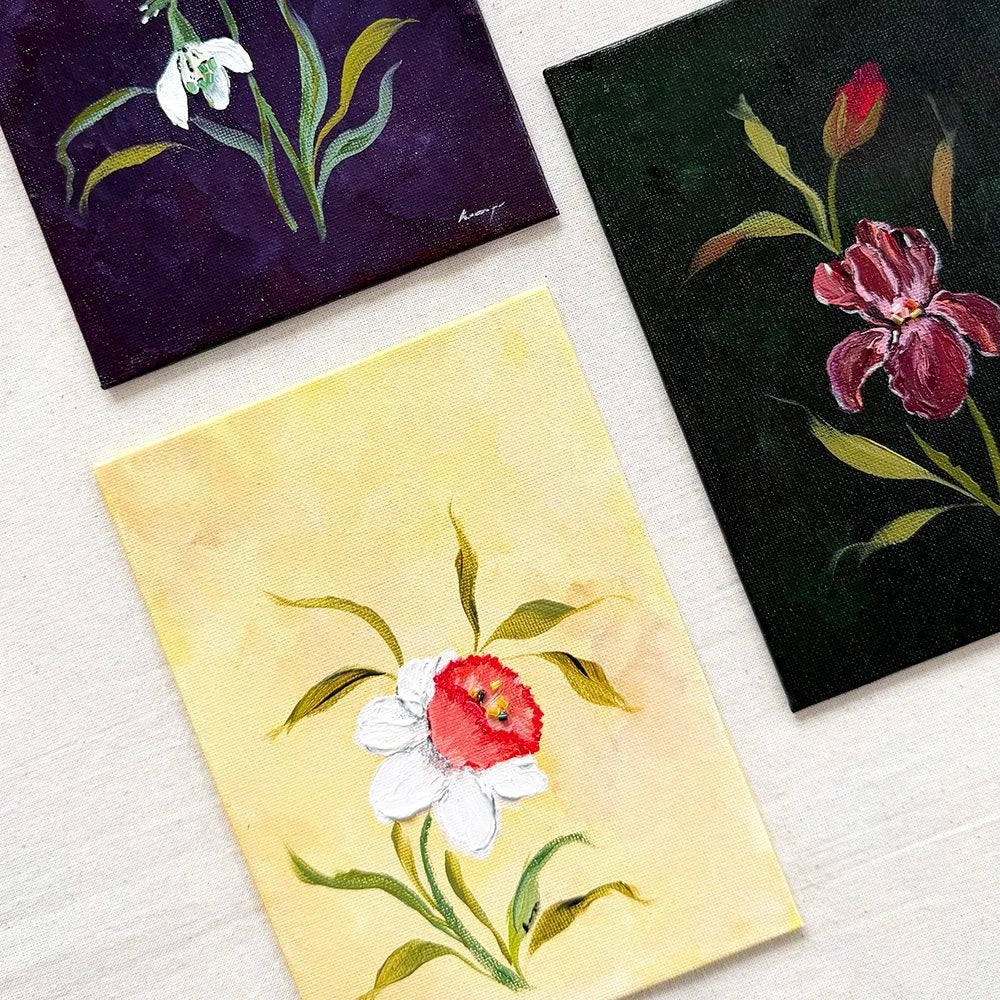 Three small square floral paintings arranged on a light textured surface. The top-left painting shows white snowdrops on a dark purple background.