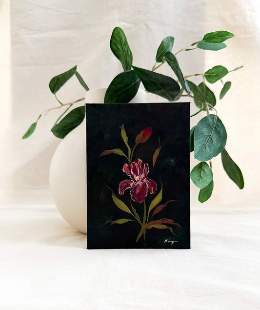 A dark green art print featuring a painted red iris flower with green and gold leaves, styled in front of a white ceramic vase with eucalyptus branches against a draped white fabric background.