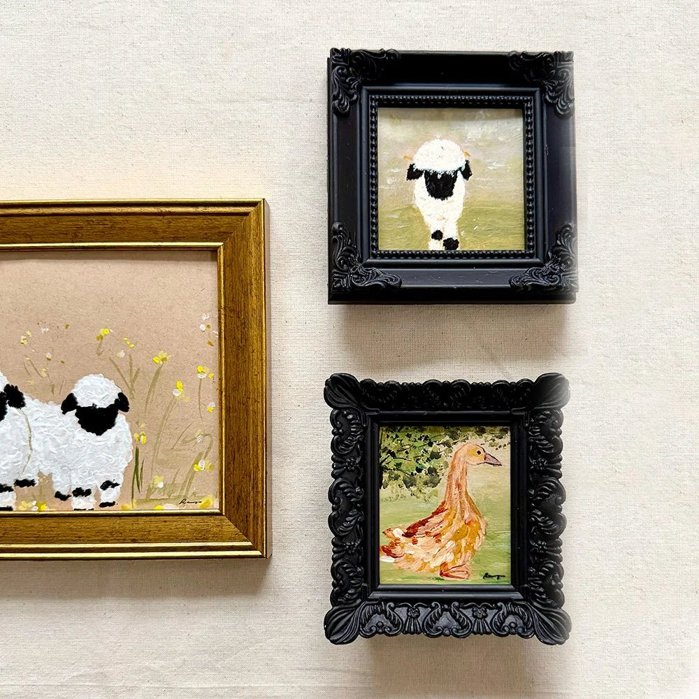 Three framed animal paintings hanging on a textured beige wall. The left frame has a gold border and shows two white sheep with black faces in a field of yellow flowers.