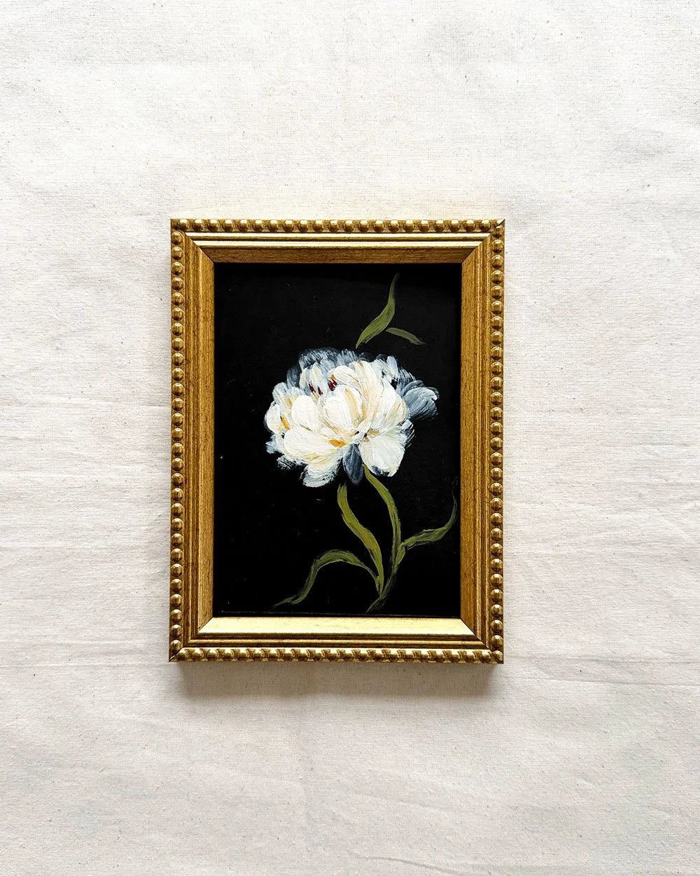 A framed floral original painting featuring a white flower with blue-gray shading and green leaves on a black background, enclosed in an ornate gold frame and mounted on a light textured wall.