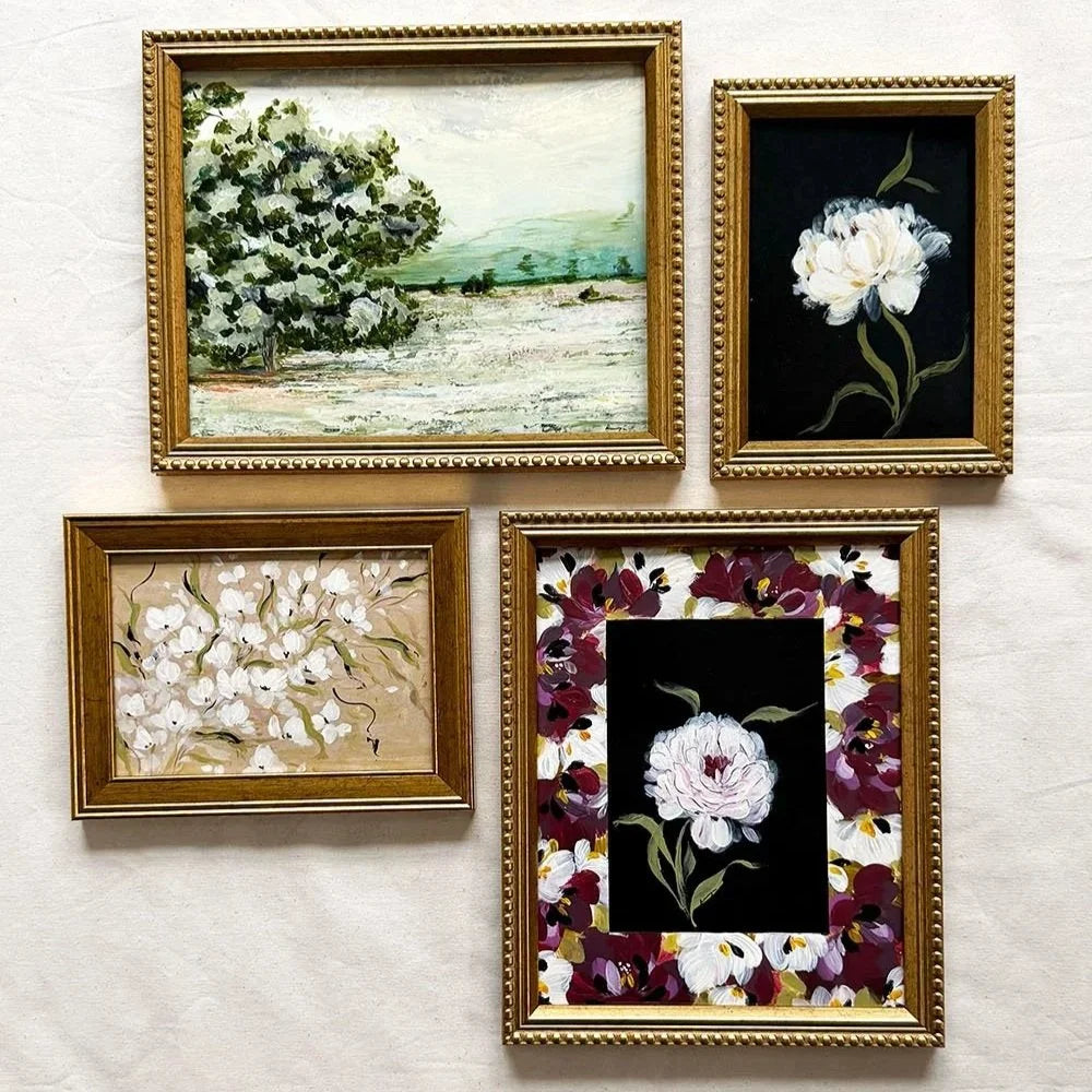 Four framed floral and landscape paintings arranged on a light fabric background. The frames are ornate gold-toned styles, with one painting depicting a tree by the water, one a white flower on black, one white flowers on beige, and one a central white flower surrounded by dark purple and white flowers on black.