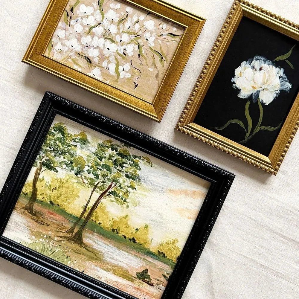 Three framed paintings arranged on a light fabric background: a black-framed landscape with trees, a gold-framed white floral painting, and a gold-framed black floral painting with a white flower.