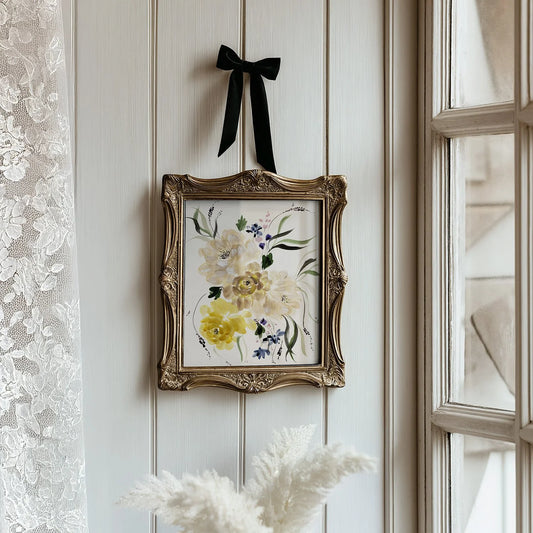 Cream and yellow floral print in an ornate gold frame with a black ribbon beside a lace-curtained window.