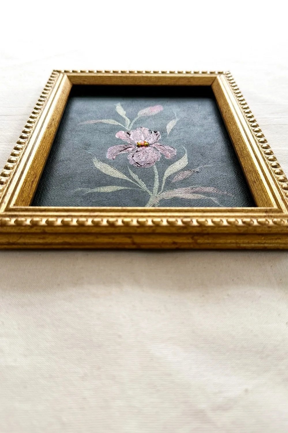 A framed floral original featuring a pink iris with green leaves on a dark gray background, enclosed in an ornate gold frame