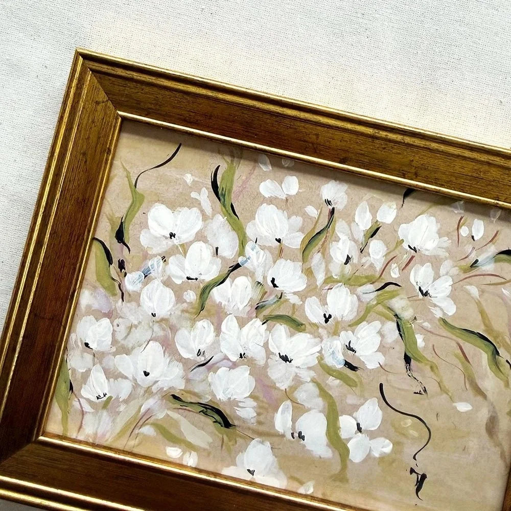 A framed painting of white magnolia blossoms with green stems on a beige background, enclosed in a gold-toned wooden frame.