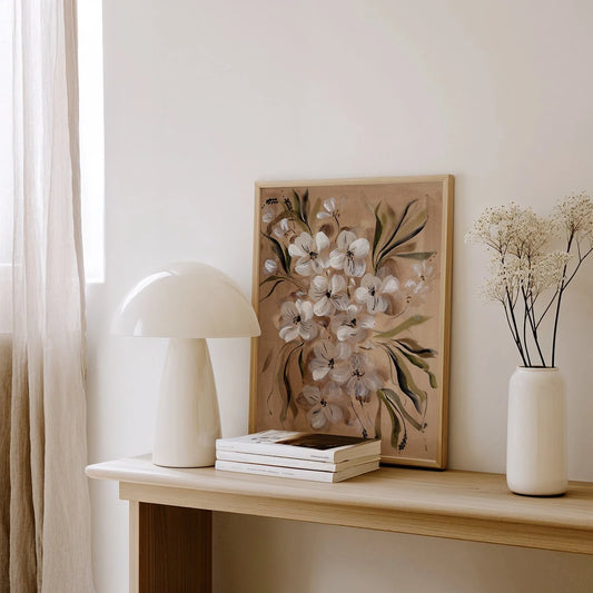 A framed floral art print with white blossoms and green leaves leaning on a light wood console table, styled with a white mushroom lamp, stacked books, and a white vase with dried flowers in a bright room.