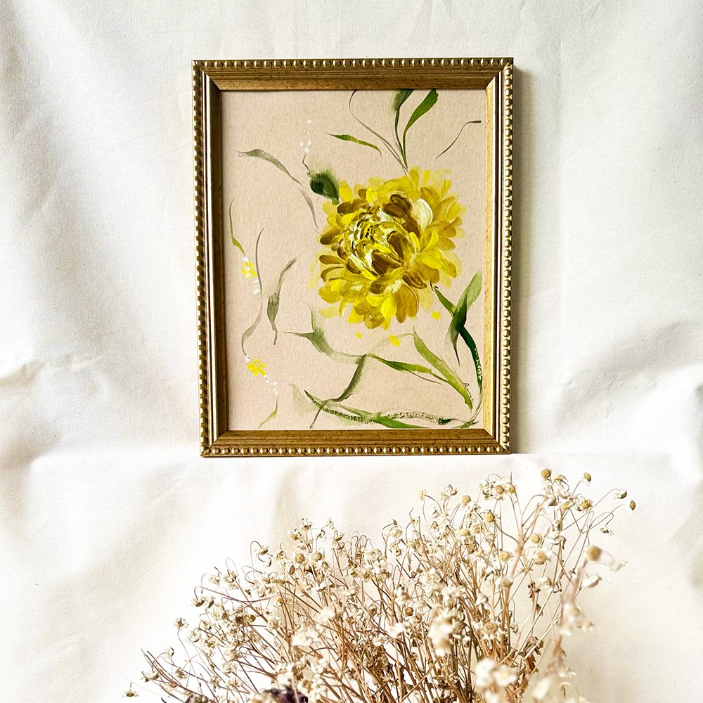 A framed floral art print featuring a yellow flower with green leaves on a textured beige background, enclosed in an ornate gold frame and styled on a white fabric backdrop with dried flowers at the bottom.