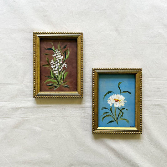 Two small framed original botanical paintings. The left frame shows white lily of the valley with green leaves on a brown background, and the right frame shows a white daisy with a yellow center on a blue background.