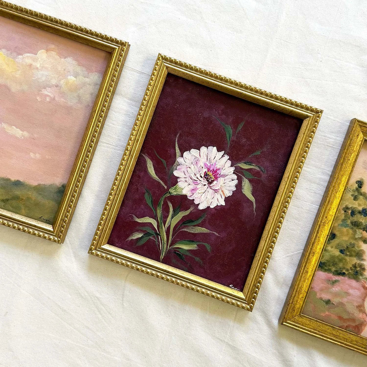 A framed floral painting featuring a white daisy with green leaves on a deep burgundy background, set in a decorative gold frame.