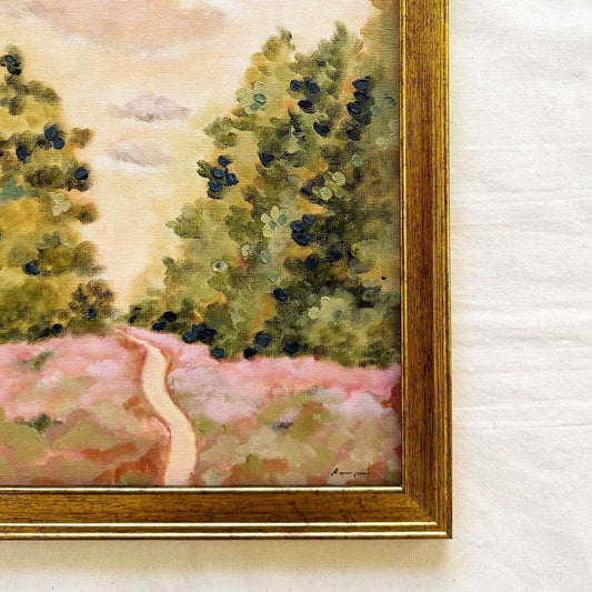 A close-up of a framed landscape original painting showing green trees, a pale pink field, and a winding path, with a gold-toned wooden frame