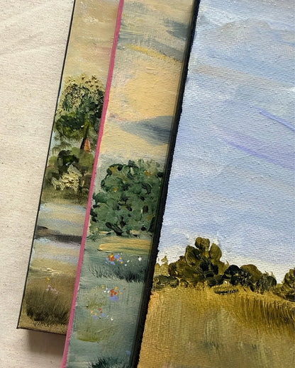 Close-up of two small landscape paintings or painted canvases leaning against a neutral surface. The left canvas shows a tree-lined waterway with green foliage, blue-gray water, and beige shore; the right canvas shows a pale blue sky over golden grass with dark tree silhouettes.