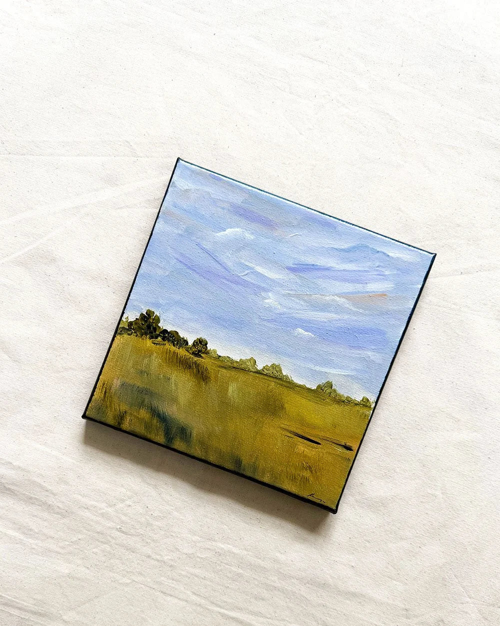 A square canvas painting of a landscape with a blue sky and green-yellow field, displayed on a textured off-white fabric background.