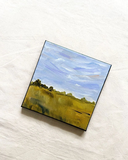 A square canvas painting of a landscape with a blue sky and green-yellow field, displayed on a textured off-white fabric background.