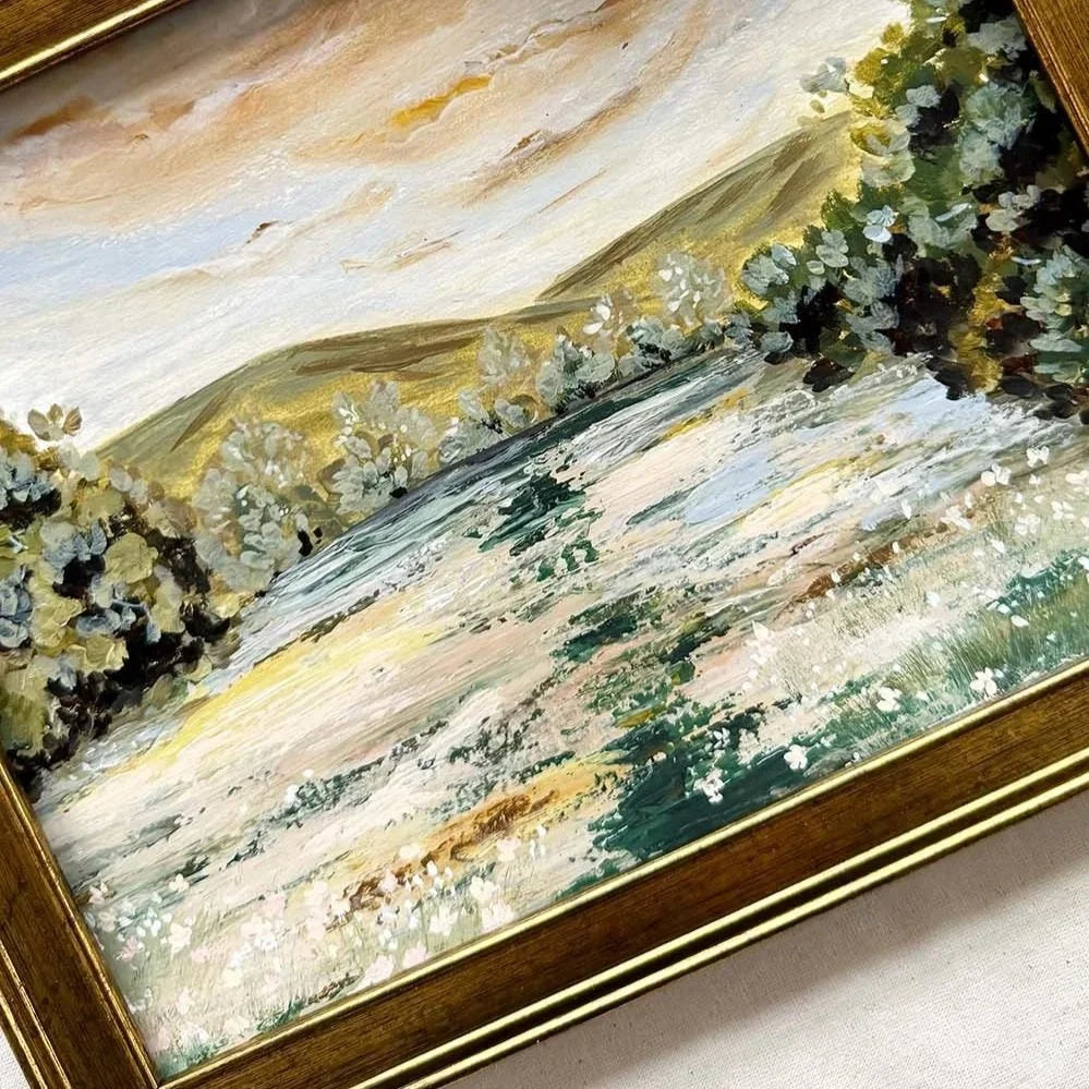 A framed landscape painting featuring rolling hills, trees, and a flowing field, rendered in soft brushstrokes of beige, green, white, and yellow, set in a gold-toned frame.
