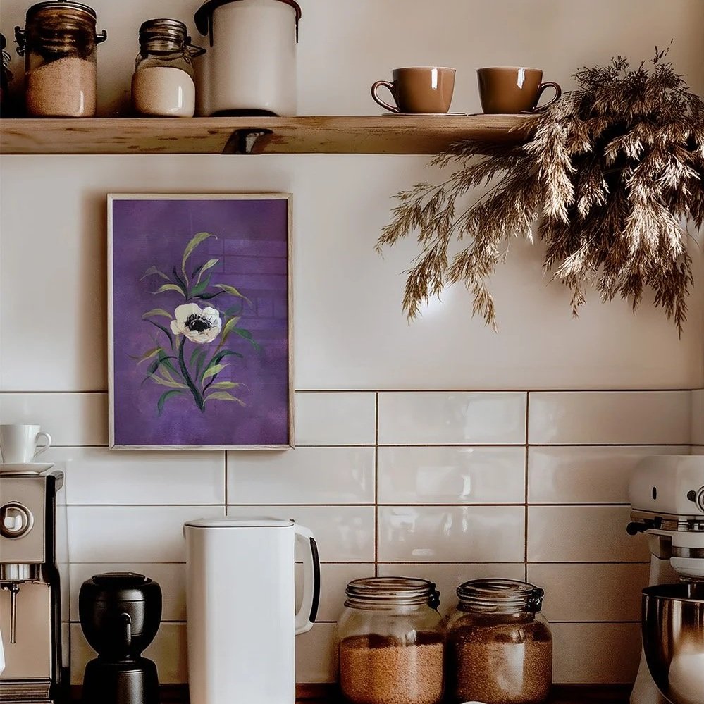 A framed art print featuring a purple background with a white anemone and green leaves, hanging on a tiled kitchen wall above a coffee station with mugs, jars, and a wooden shelf with dried grasses.