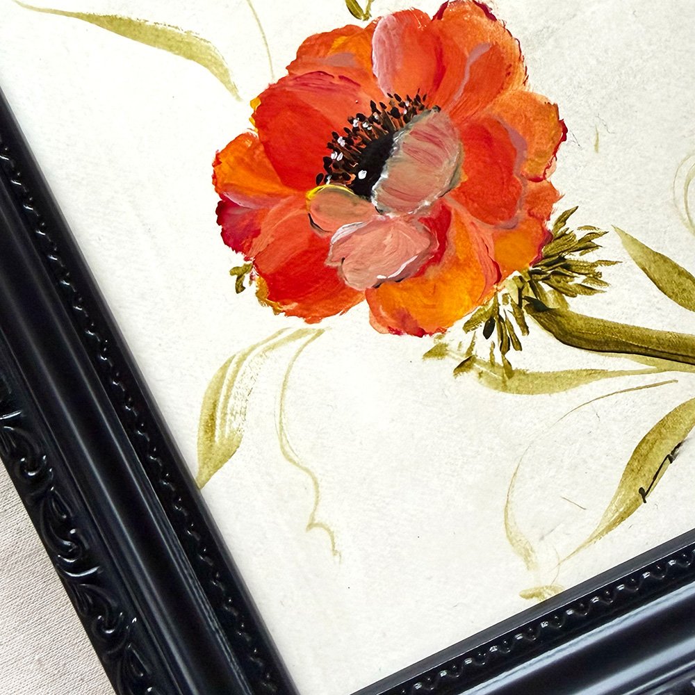 A close-up of framed floral wall art showing a large orange-red anemone flower with green leaves on an off-white background, with part of a black ornate frame visible.