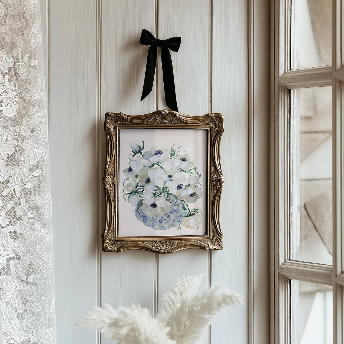 White anemone vase print in an ornate gold frame suspended by a black ribbon beside a window.
