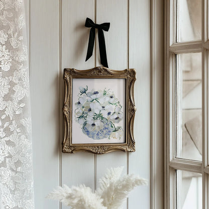 White anemone vase print in an ornate gold frame suspended by a black ribbon beside a window.