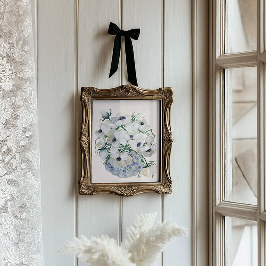 White anemone vase print in an ornate gold frame suspended by a black ribbon beside a window.