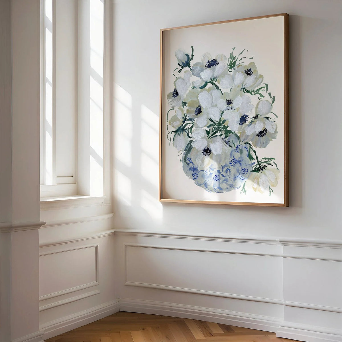 Large white anemone vase print in a gold frame on a paneled wall beside a sunlit window.