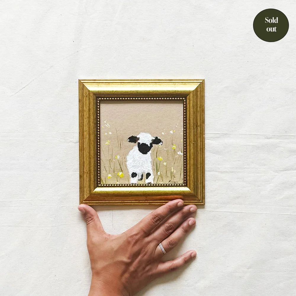 A small framed artwork showing a white sheep with black facial features standing in a field of yellow flowers on a beige background, held by a person's hand against a white textured backdrop. A dark green 'Sold out' badge appears in the top right corner.