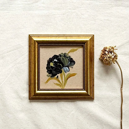 A framed botanical art original featuring a dark flower with green and blue accents on a beige background, enclosed in a gold frame