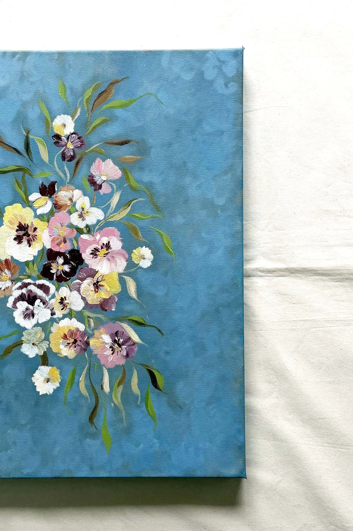 A canvas painting featuring a bouquet of pansies in soft pink, yellow, white, and purple tones on a textured blue background. Perfect as a dinning room decor by Ramya Rajiv