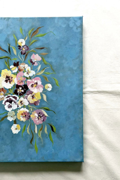 A canvas painting featuring a bouquet of pansies in soft pink, yellow, white, and purple tones on a textured blue background. Perfect as a dinning room decor by Ramya Rajiv