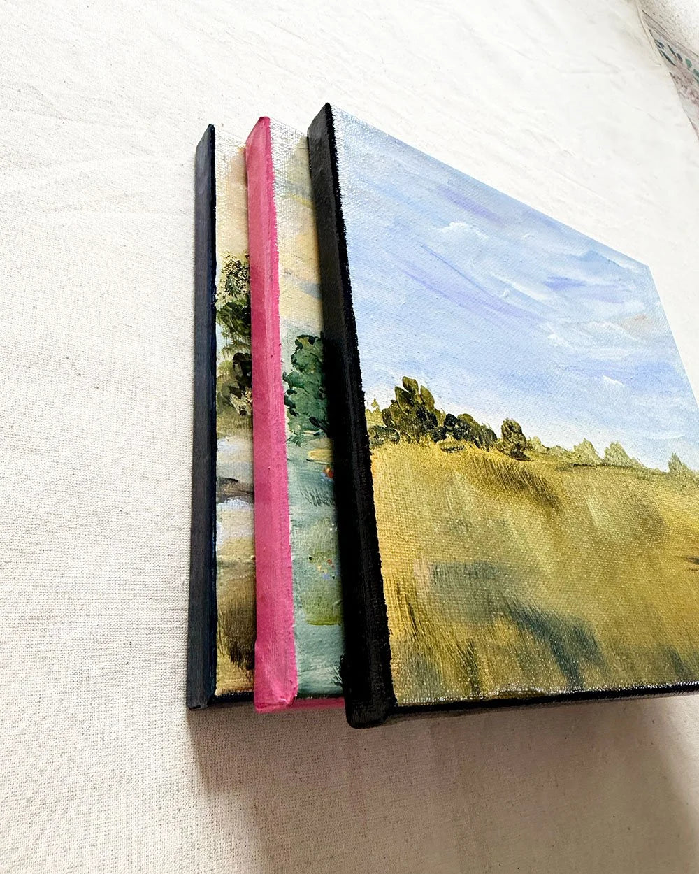 Three small canvas paintings fanned out on a light fabric surface. The front canvas shows a landscape with a blue sky, white clouds, and a yellow-green field with dark trees.