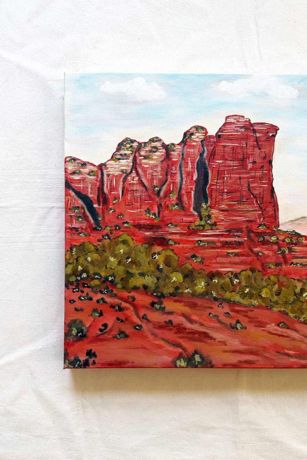 A original canvas painting of a red rock desert landscape, of Sedona Coffee Pot Rock featuring tall red sandstone cliffs, green shrubs, and a light blue sky, displayed on a white fabric background.