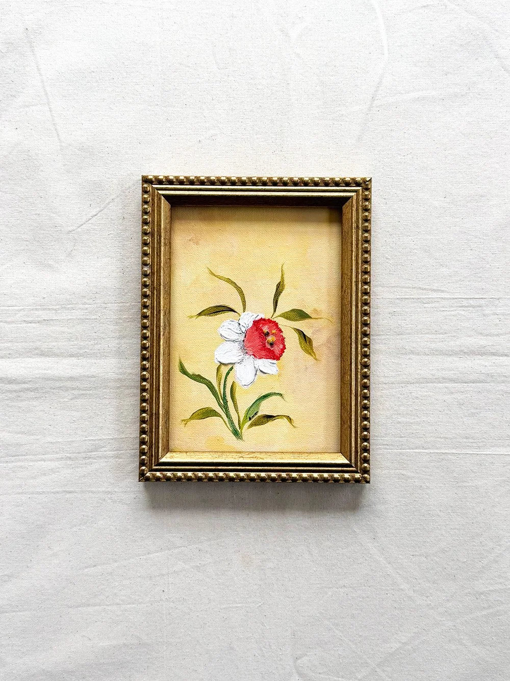 A small framed original floral painting featuring a Daffodil with white petals and green leaves on a yellow background, enclosed in an ornate gold frame 