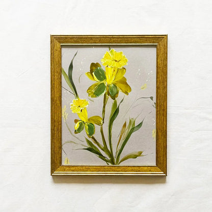 A framed painting of yellow flowers with green leaves on a light gray background, enclosed in a gold frame and mounted on a textured white wall.