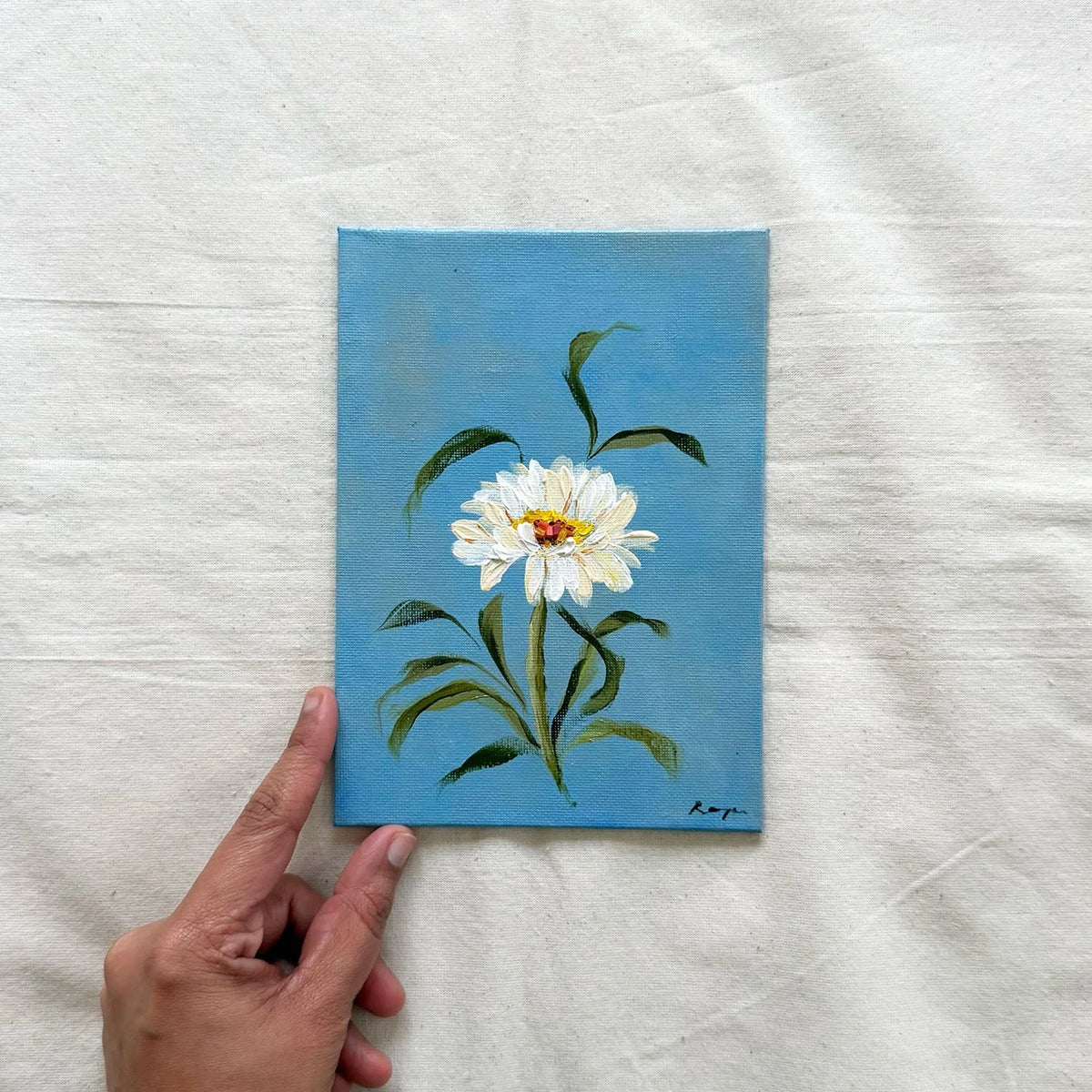 A original painting of a white daisy with green leaves on a blue background. April flower of the month