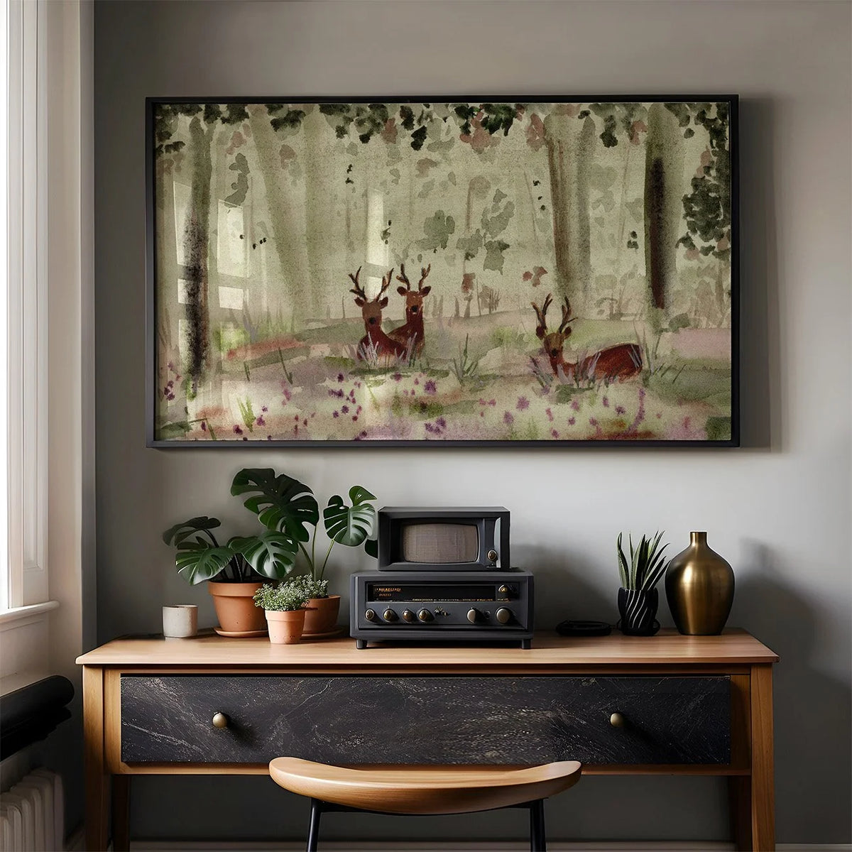 A framed art print depicting a misty forest with deer, displayed on a gray wall above a wooden console table with plants and a vintage radio.