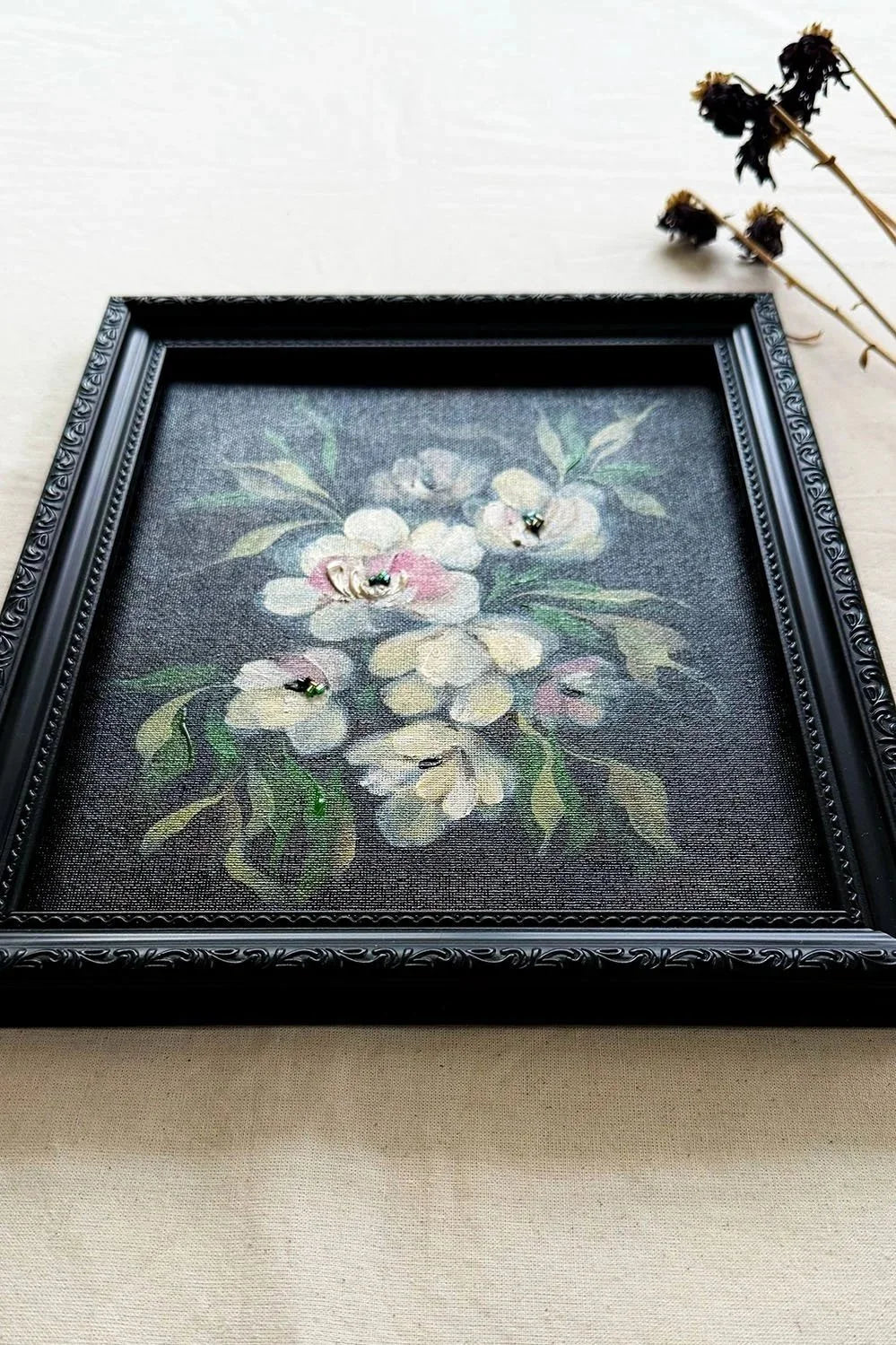 A framed floral original artwork featuring white and pink flowers with green leaves on a dark gray background, enclosed in an ornate black frame and styled on a light beige fabric surface with dried flowers in the corner.