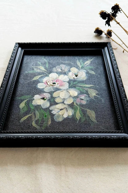 A framed floral original artwork featuring white and pink flowers with green leaves on a dark gray background, enclosed in an ornate black frame and styled on a light beige fabric surface with dried flowers in the corner.