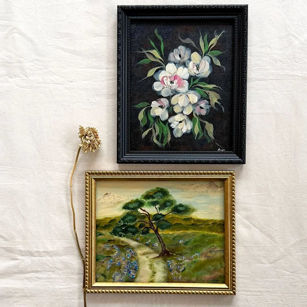 Two framed paintings displayed on a white fabric background. The top frame contains a black floral painting with white blossoms and green leaves in a black frame.