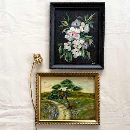 Two framed paintings displayed on a white fabric background. The top frame contains a black floral painting with white blossoms and green leaves in a black frame.