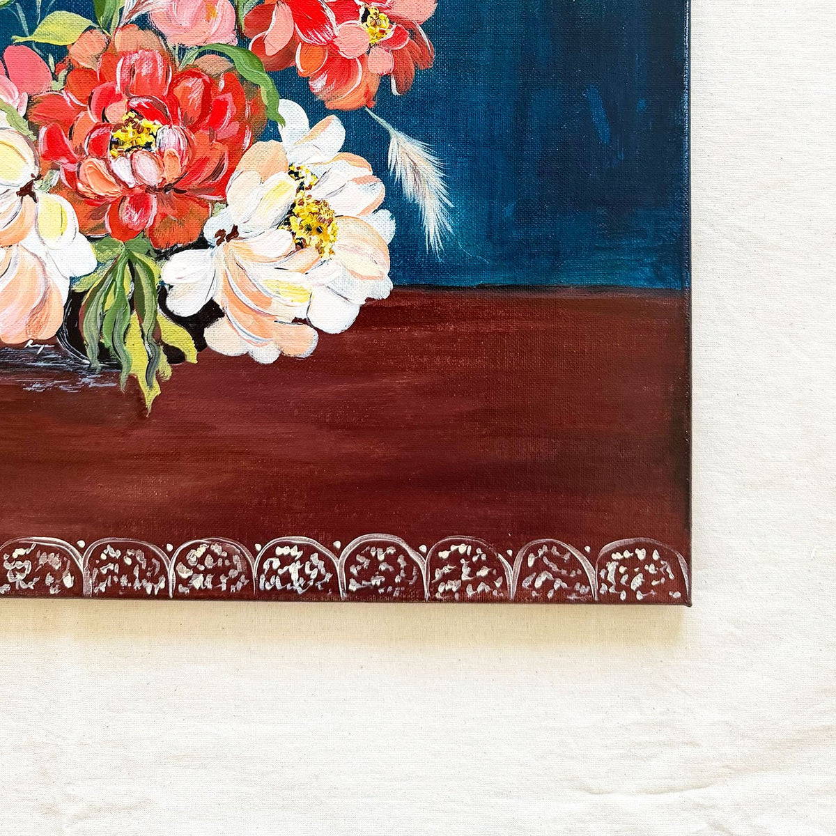 A close-up of a painted floral artwork showing red, coral, and white flowers with green leaves against a dark blue and deep brown background, mounted on a textured off-white wall.
