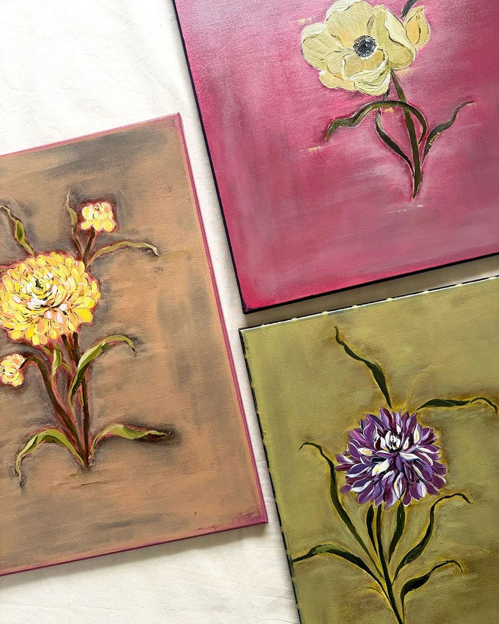 Three square floral paintings arranged on a white fabric background, each showing a single flower with textured brushstrokes: a yellow flower on brown, a pale yellow flower on pink, and a purple flower on olive green.