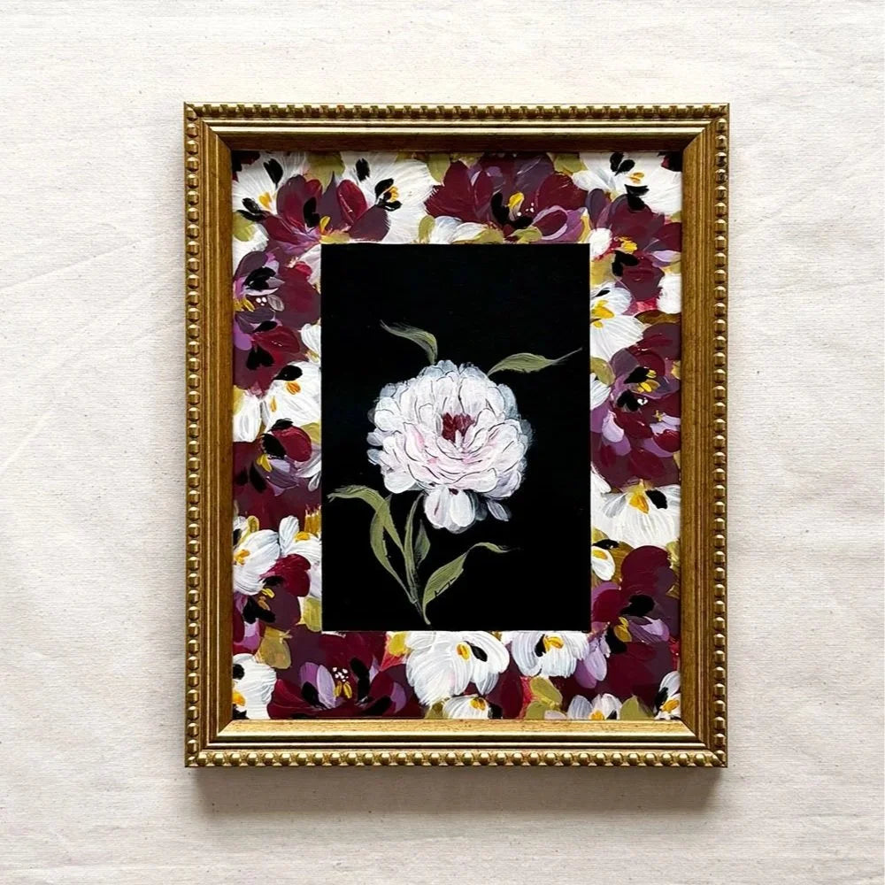 A framed floral art print featuring a central white peony on a black background, surrounded by a gold frame and a border of burgundy, white, and gold flowers