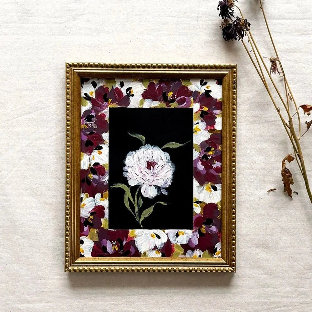 A framed floral art original featuring a central white flower on a black background, surrounded by a gold ornate frame and a border of burgundy, white, and yellow flowers