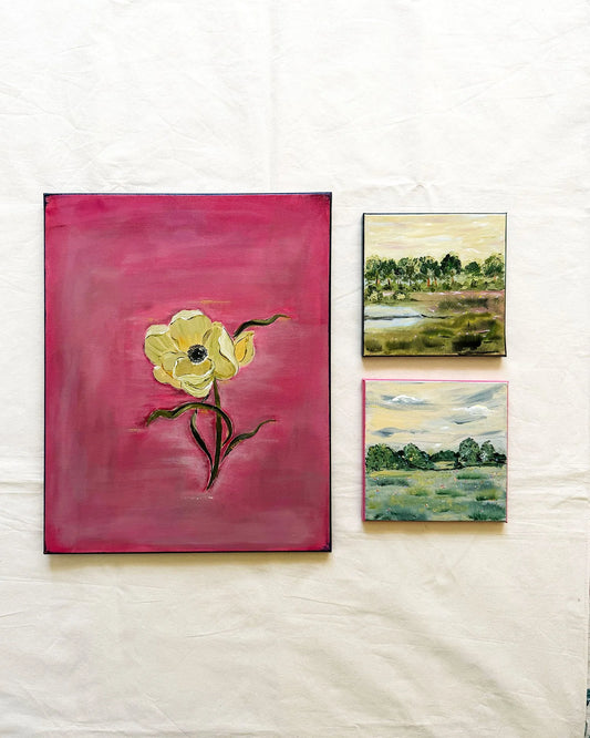 Three paintings displayed on a white fabric background: a large vertical canvas featuring a yellow flower on a pink background, and two smaller landscape paintings with green, blue, and yellow tones.