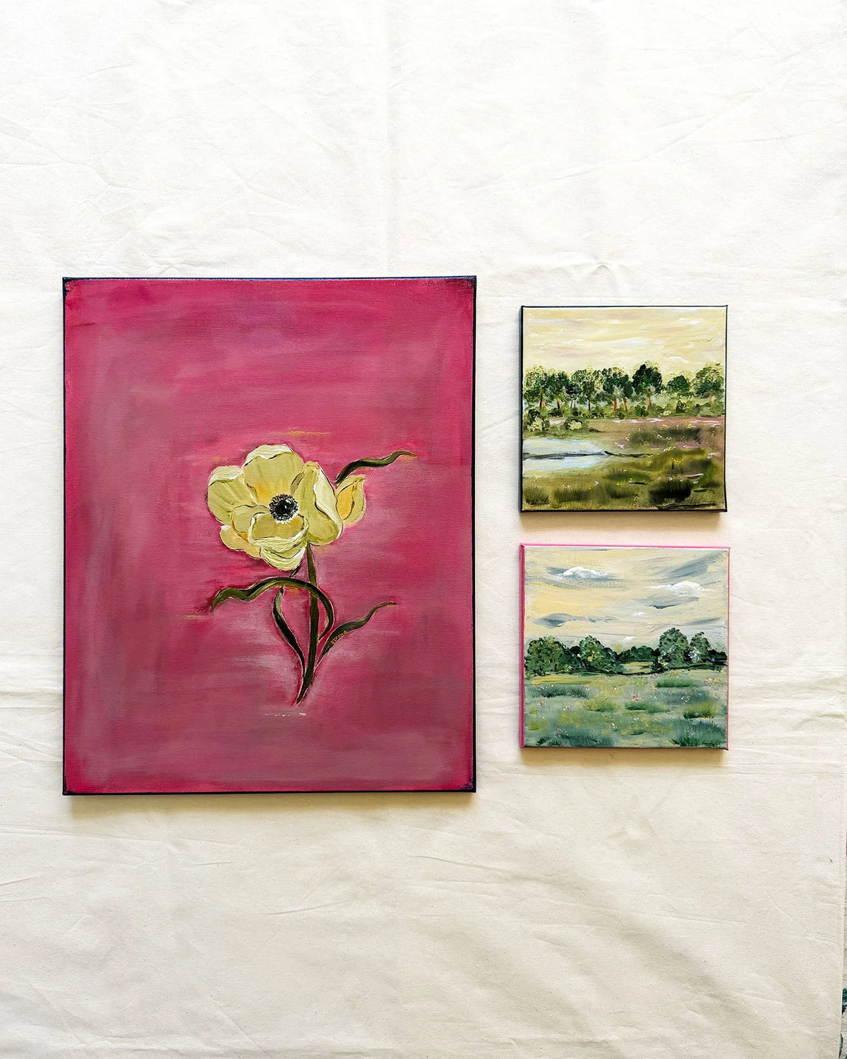Three paintings displayed on a white fabric background: a large vertical canvas featuring a yellow flower on a pink background, and two smaller landscape paintings with green, blue, and yellow tones.