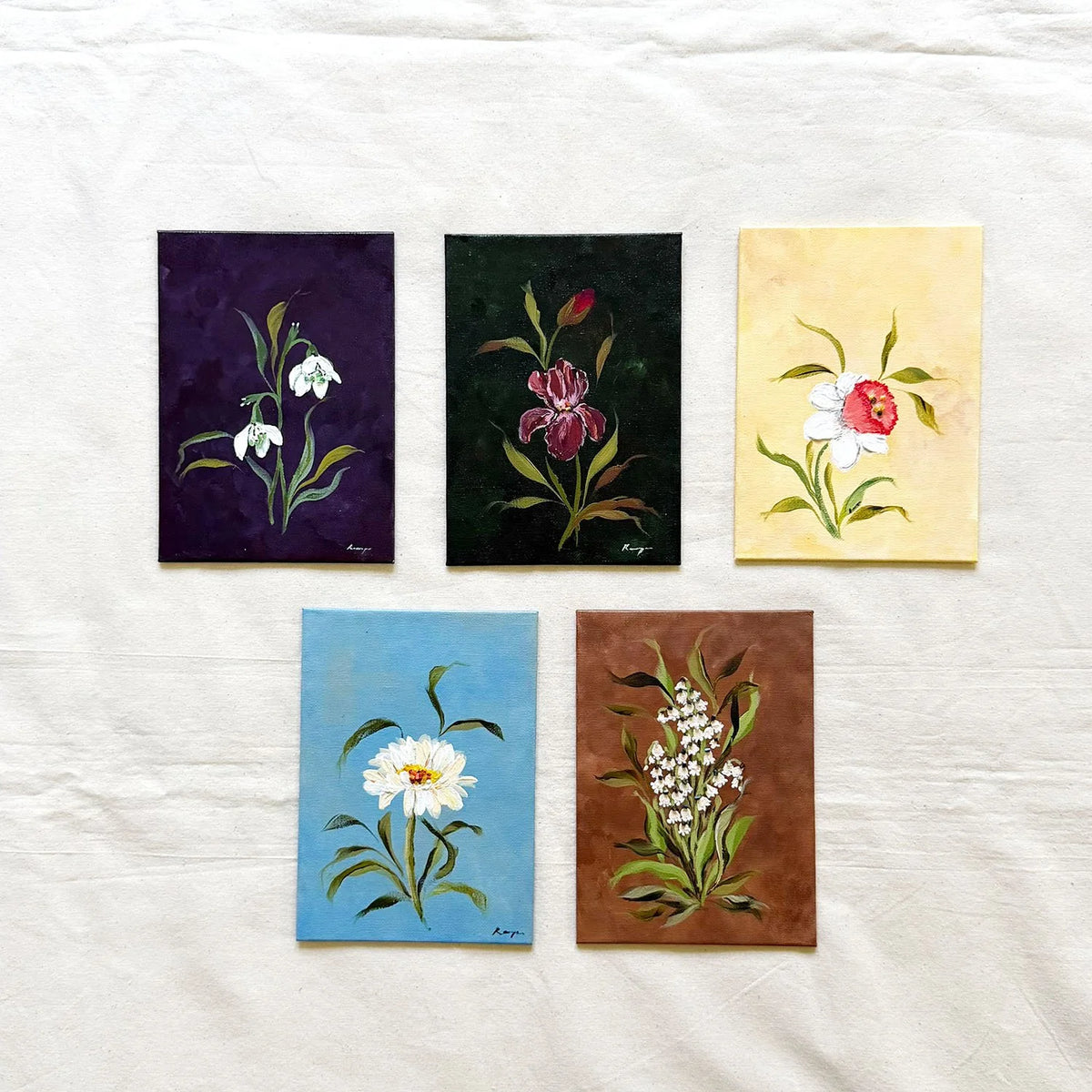 Five small original floral paintings. The paintings feature white snowdrops on a dark purple background, a pink iris on a very dark green background, daffodil on a pale yellow background, a white daisy on a light blue background, and white lily of the valley on a brown background.