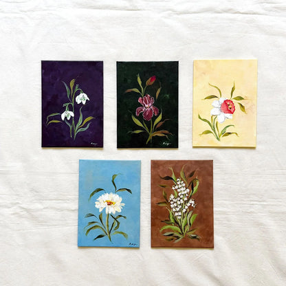 Five small original floral paintings. The paintings feature white snowdrops on a dark purple background, a pink iris on a very dark green background, daffodil on a pale yellow background, a white daisy on a light blue background, and white lily of the valley on a brown background.