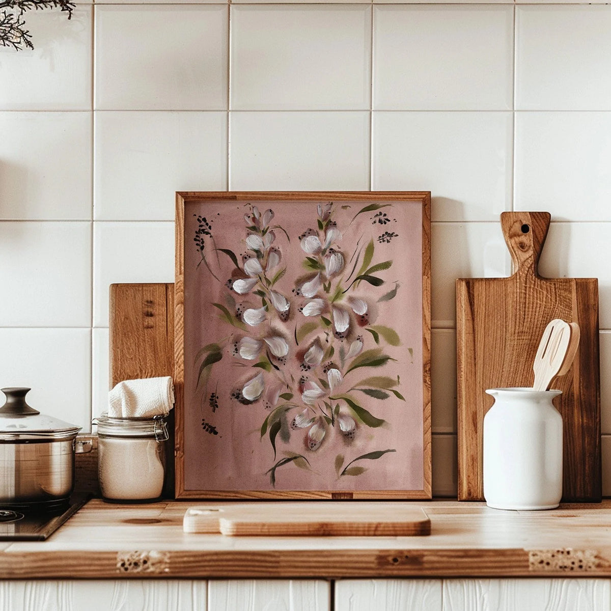 A framed botanical art print featuring white flowers and green leaves on a dusty pink background, styled on a wooden kitchen counter with cutting boards, a white pitcher, and tiled walls behind.