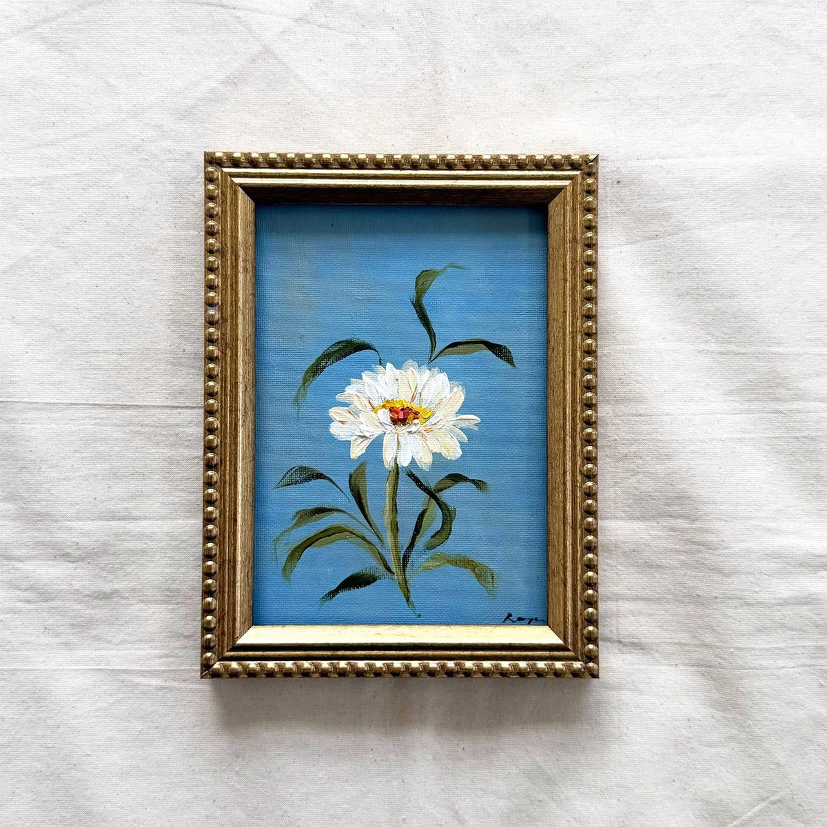 A framed original painting of a white flower with green leaves on a blue background, set in an ornate gold frame 